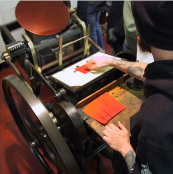 Vintage letterpress machine mid-print with a craftsperson's hands guiding orange stock.